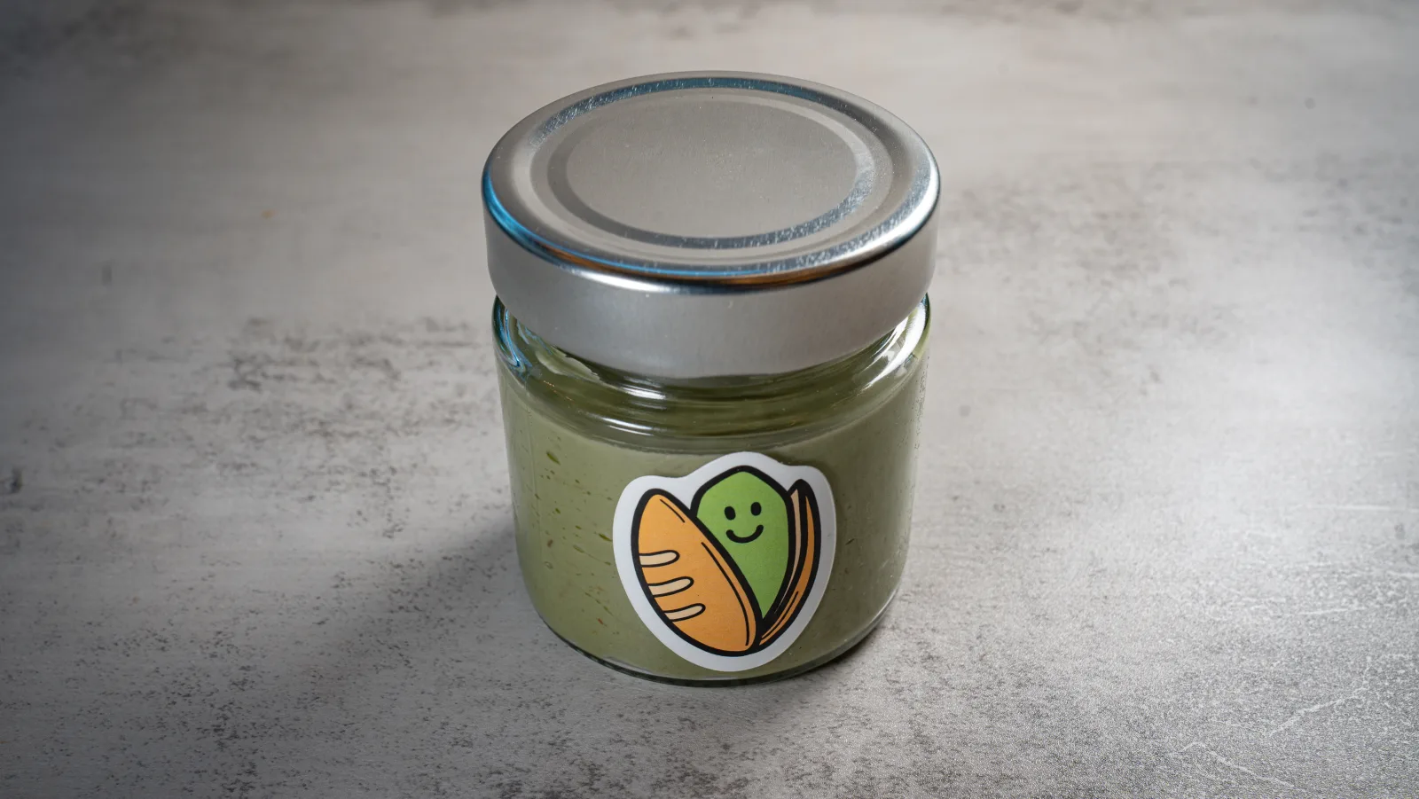 A jar of Pistacchio's own pistachio cream on grey concrete, the colour a muted olive green
